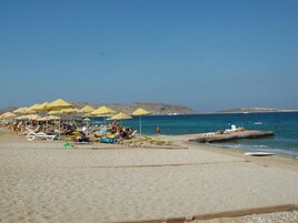 Una playa con tumbonas y sombrillas, un muelle que se adentra en el mar y colinas rocosas al fondo.