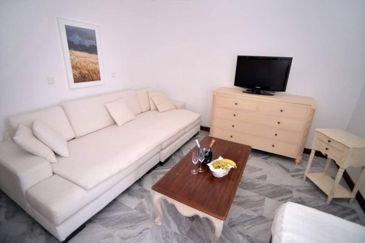 A living room with a beige sofa, a wooden coffee table with a bottle and glasses, a TV on a dresser, and a framed picture on the wall.