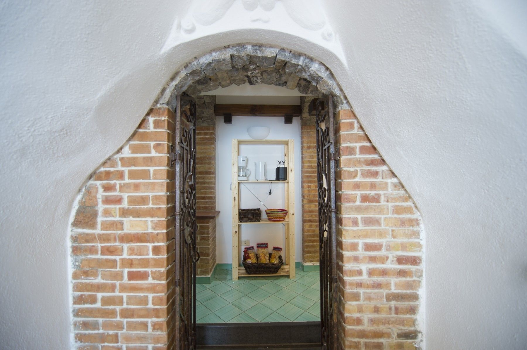 A narrow brick archway leading to a room with a wooden door, a shelf with kitchen items, and a basket of snacks.