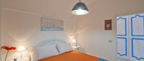 A bedroom with a bed, a nightstand, a lamp, a painting, and a door with blue trim.