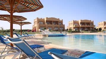 A poolside area with lounge chairs and umbrellas, a clear blue sky, and a building with balconies in the background.
