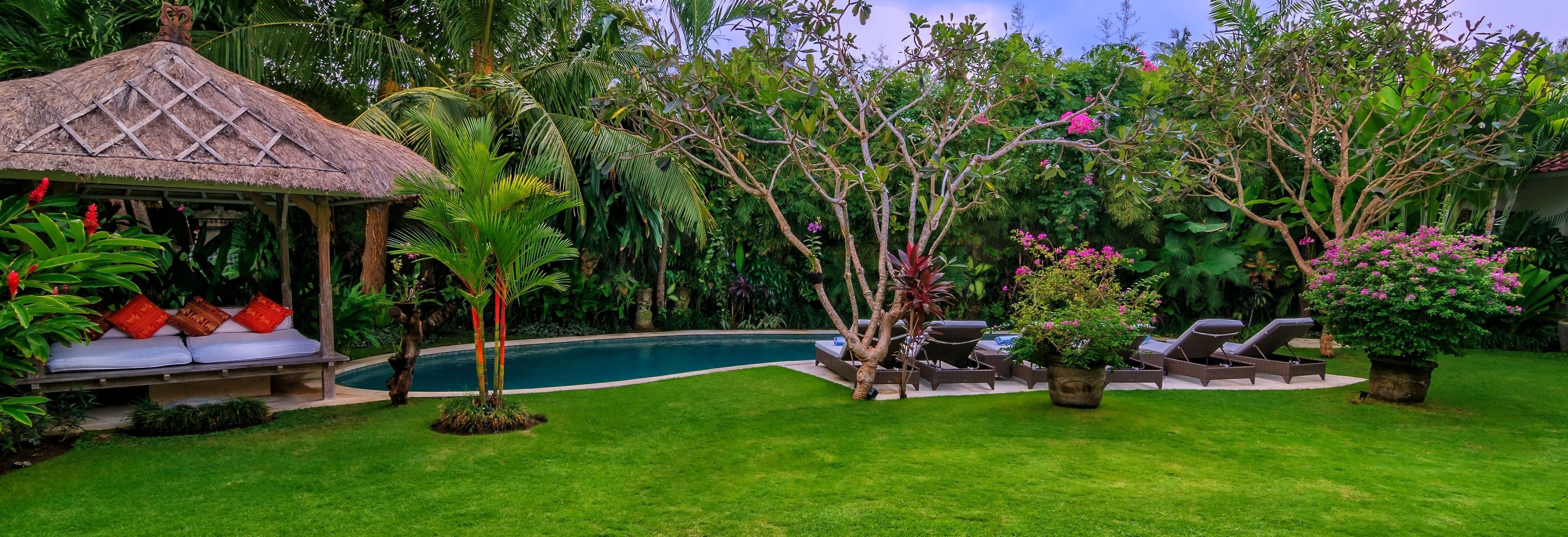 A poolside area with lounge chairs, a thatched-roof gazebo, and lush greenery.
