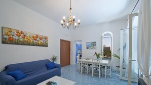 A living room with a blue sofa, a white coffee table, and a chandelier. There is a dining area with a white table and chairs. - Casa Agata (Praiano)