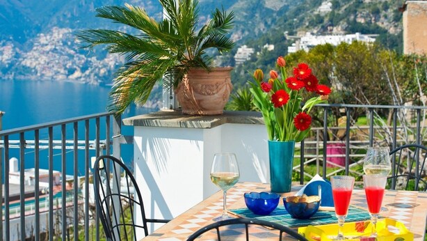 A table with a potted plant, red flowers, and two glasses of red drink, overlooking a scenic coastal view. - Casa Agata (Praiano)