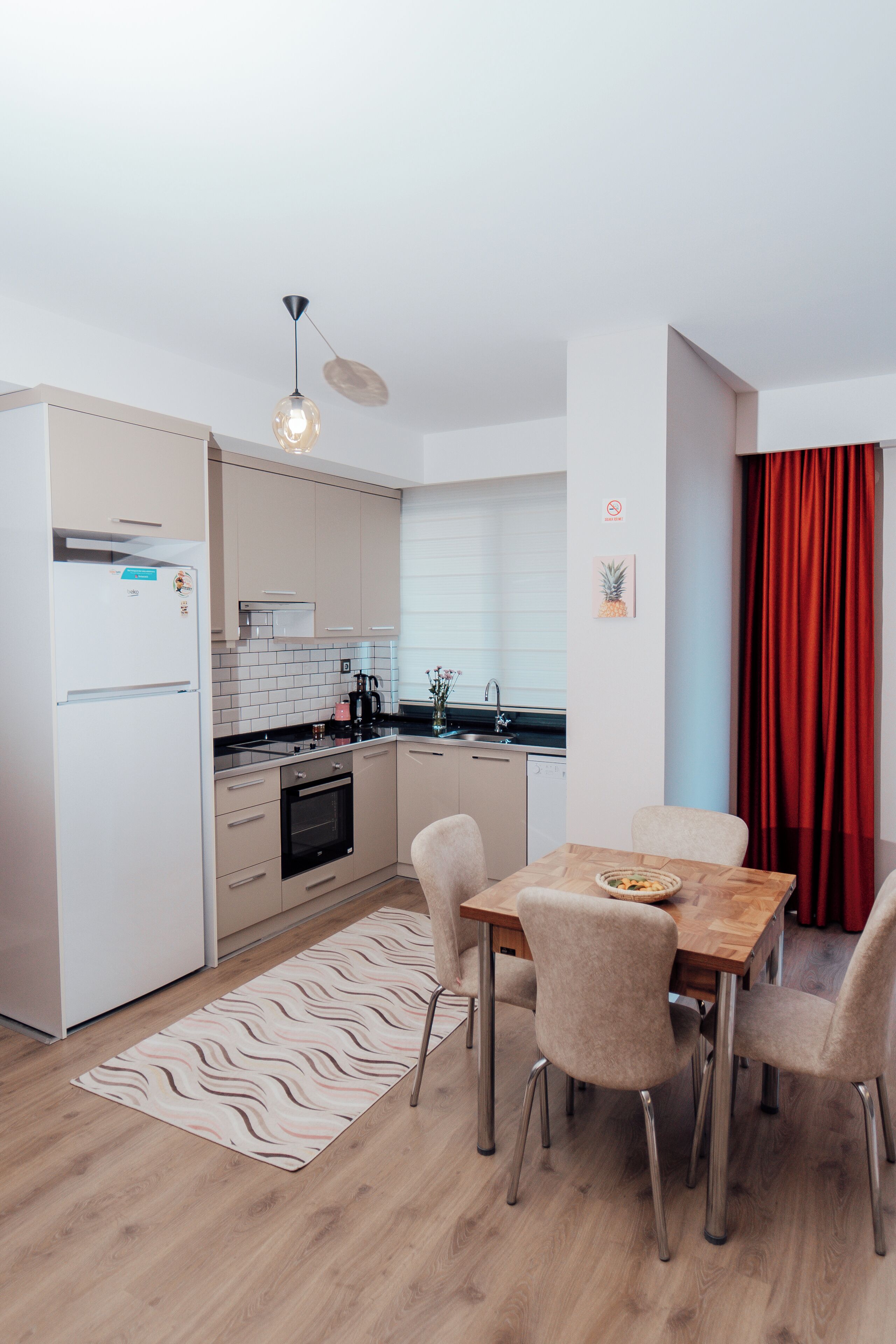 A modern kitchen with a dining area, featuring a wooden table, beige chairs, and a red curtain.