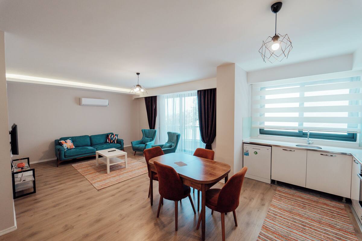 Un salon moderne comprenant un canapé, des fauteuils, une table à manger et un coin cuisine avec une fenêtre et des rideaux.