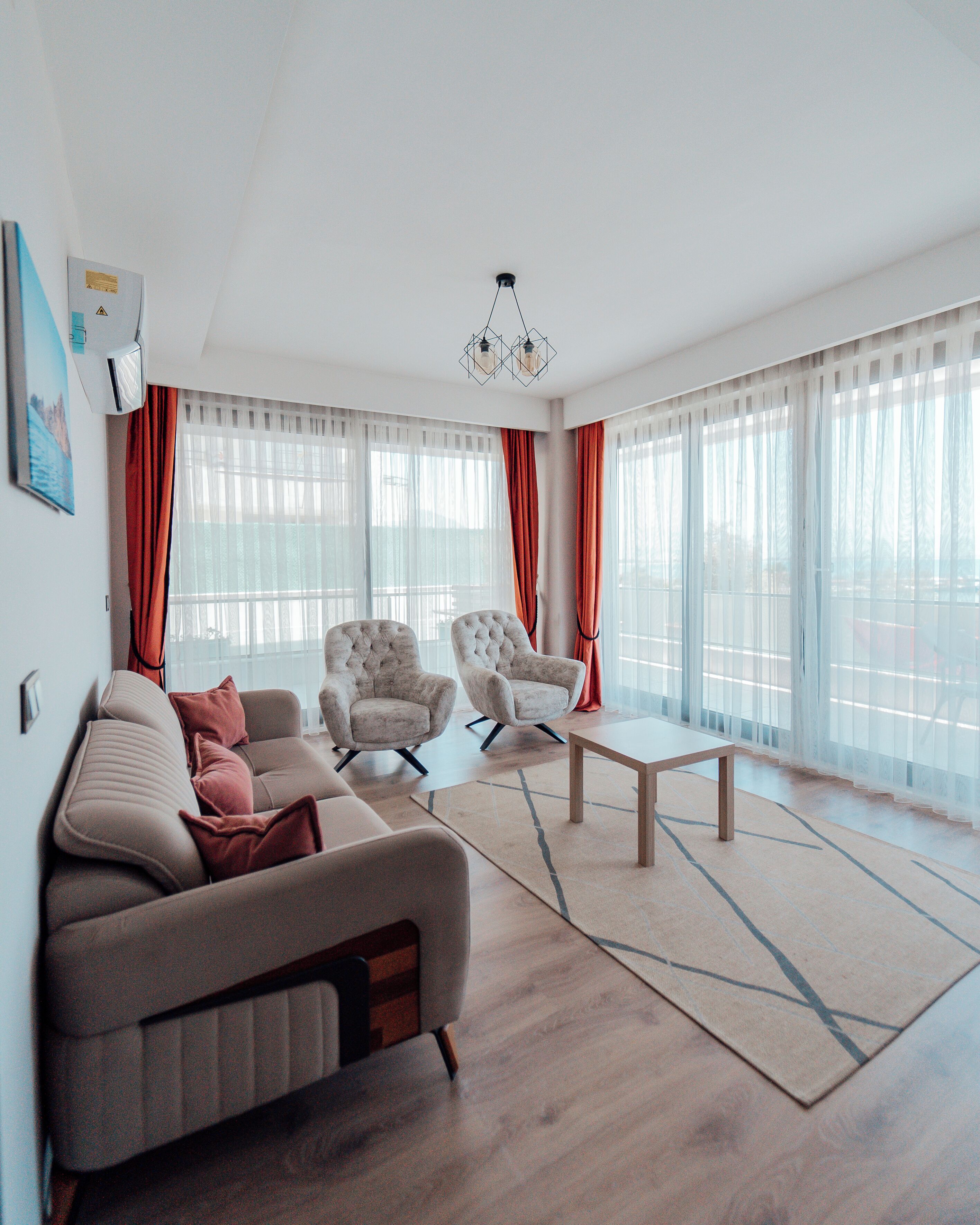 Ein modernes Wohnzimmer mit einer Couch, zwei Sesseln, einem Couchtisch und großen Fenstern mit durchsichtigen Vorhängen.