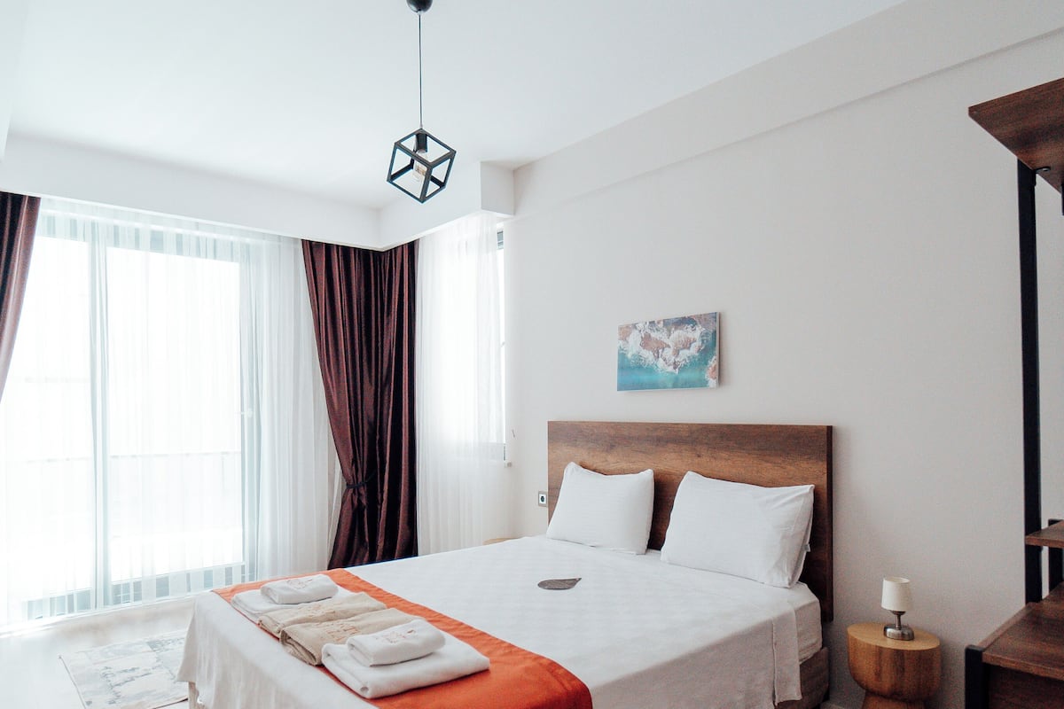 Une chambre moderne avec un grand lit, une tête de lit en bois, une petite table de chevet avec une lampe, et une vue sur l’extérieur grâce à de grandes fenêtres.