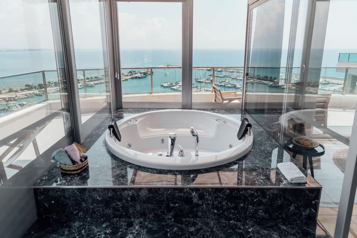 Une salle de bain moderne avec une grande baignoire, un balcon vitré offrant une vue sur la mer et les bateaux, et un comptoir en marbre.