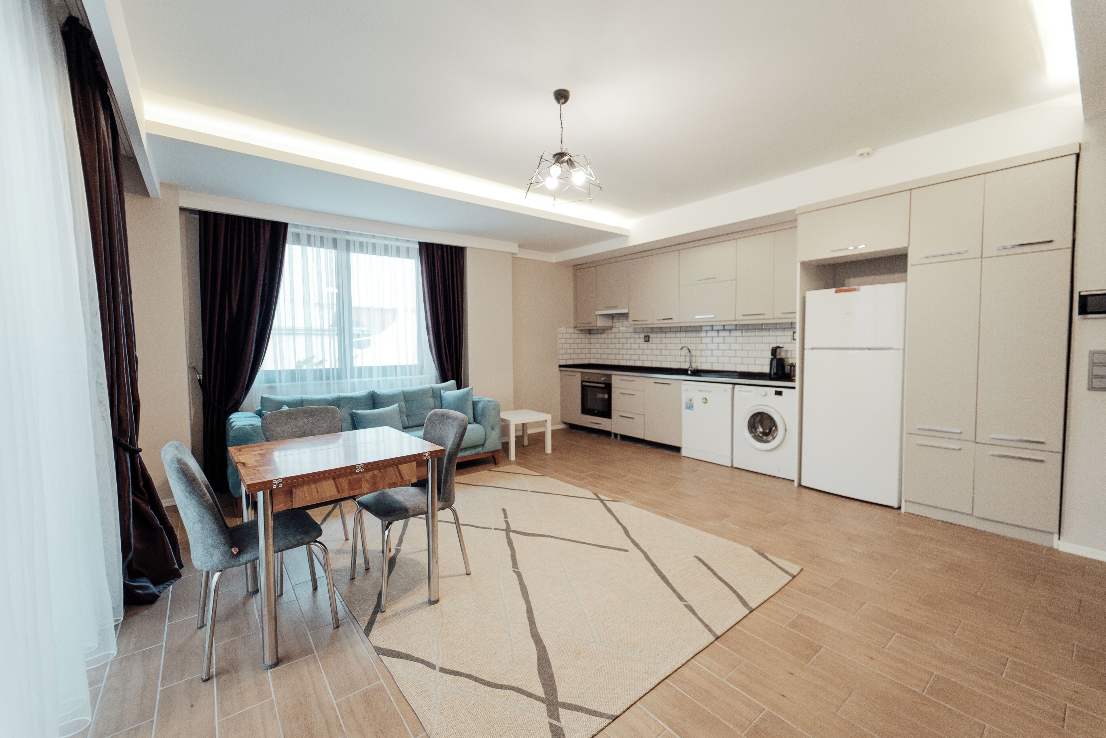 A modern kitchen with white cabinets, a built-in oven, and a refrigerator. There is a dining area with a wooden table and chairs.