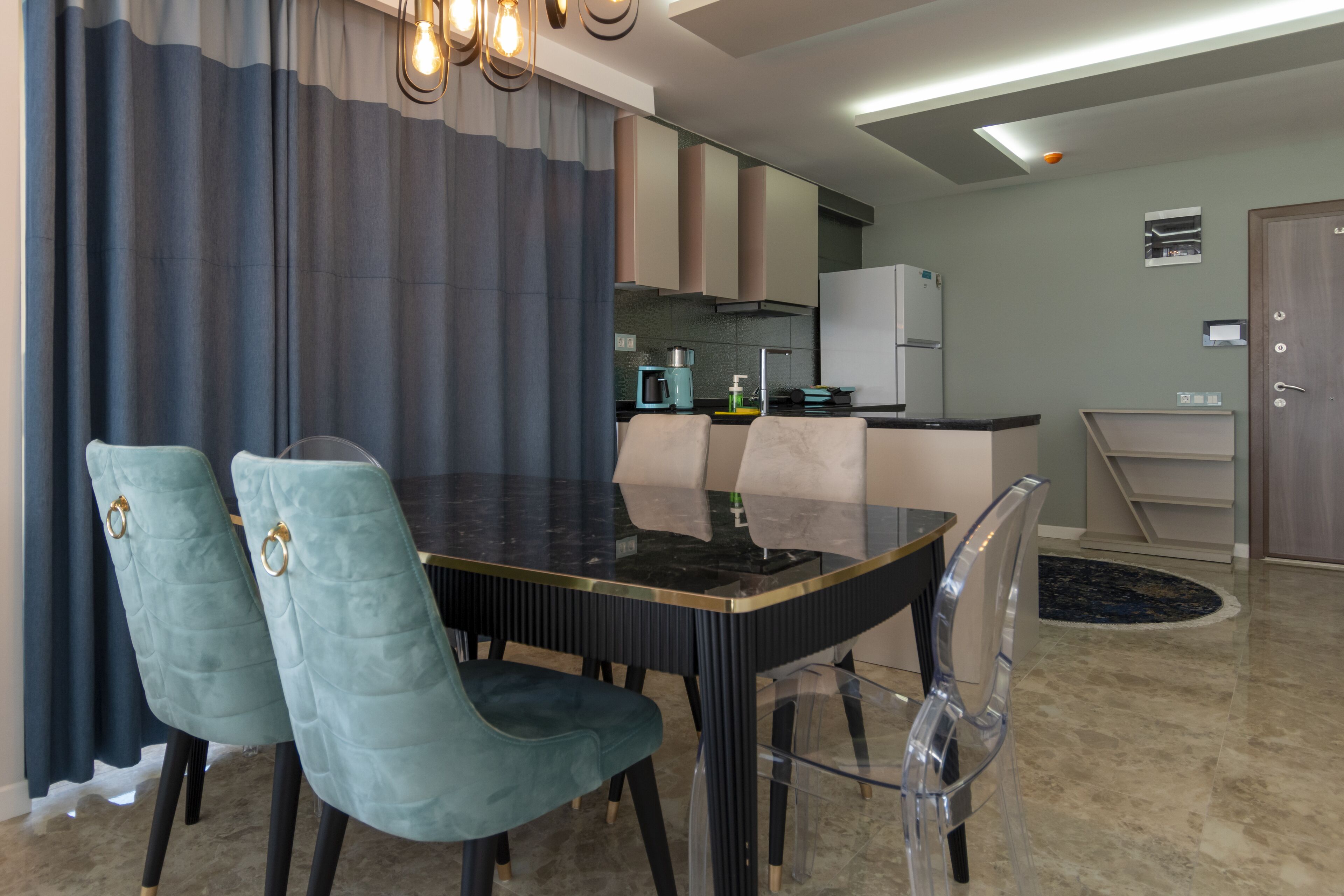 Un comedor moderno con una mesa de vidrio, dos sillas tapizadas, una cocina al fondo y una puerta a la derecha.