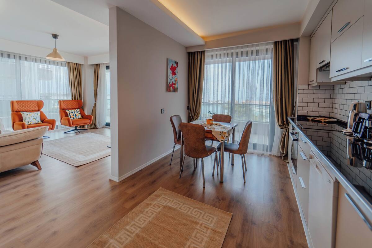 Un salon moderne comprenant un canapé, des fauteuils, une table à manger avec des chaises, un coin cuisine avec une cuisinière et des placards, ainsi que de grandes fenêtres avec des rideaux.