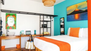 A hotel room with a bed, a desk with a laptop, a shelf with a safe, and a painting of a volcano. - Bird of Paradise Hotel (Jacó)