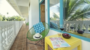 A balcony with a chair, a table with a magazine, and a window with a view of palm trees. - Bird of Paradise Hotel (Jacó)