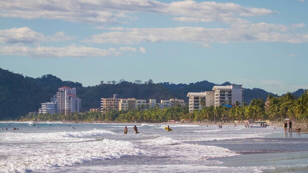 A beach with waves, people, and buildings in the background. - Bird of Paradise Hotel (Jacó)