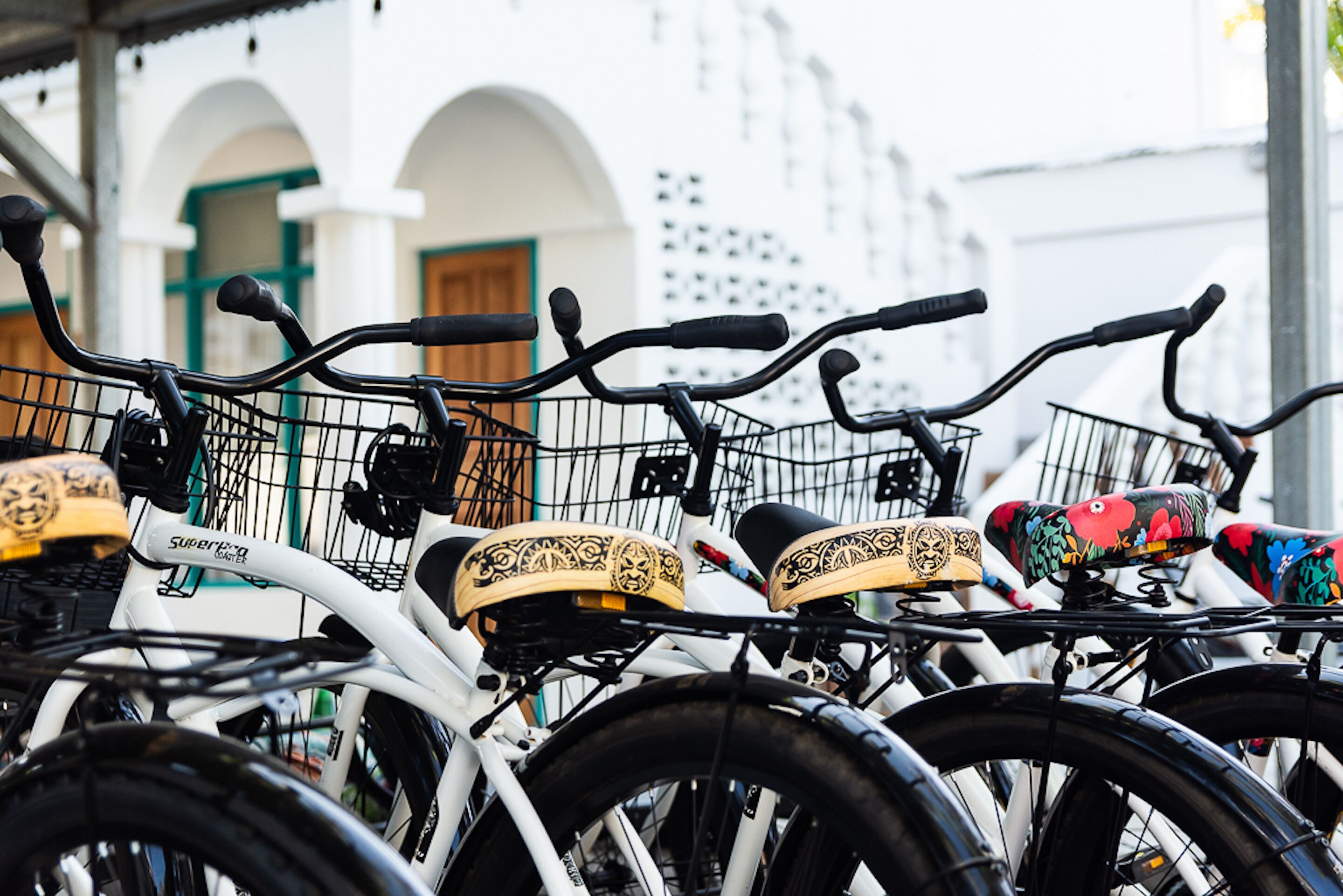 A row of bicycles with various seat designs, parked in front of a building with white walls and green accents.