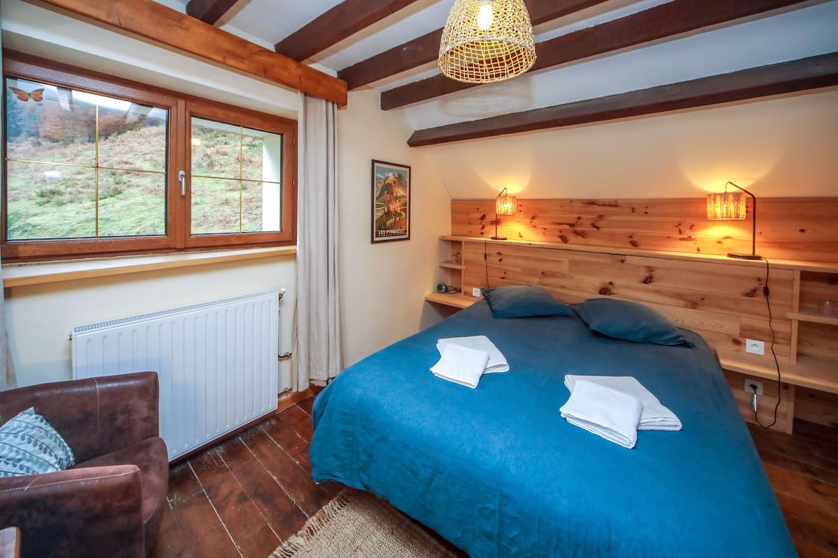 Ett sovrum med en stor säng, en sänggavel i trä, sänglampor, ett element och ett fönster med utsikt över omgivningen.