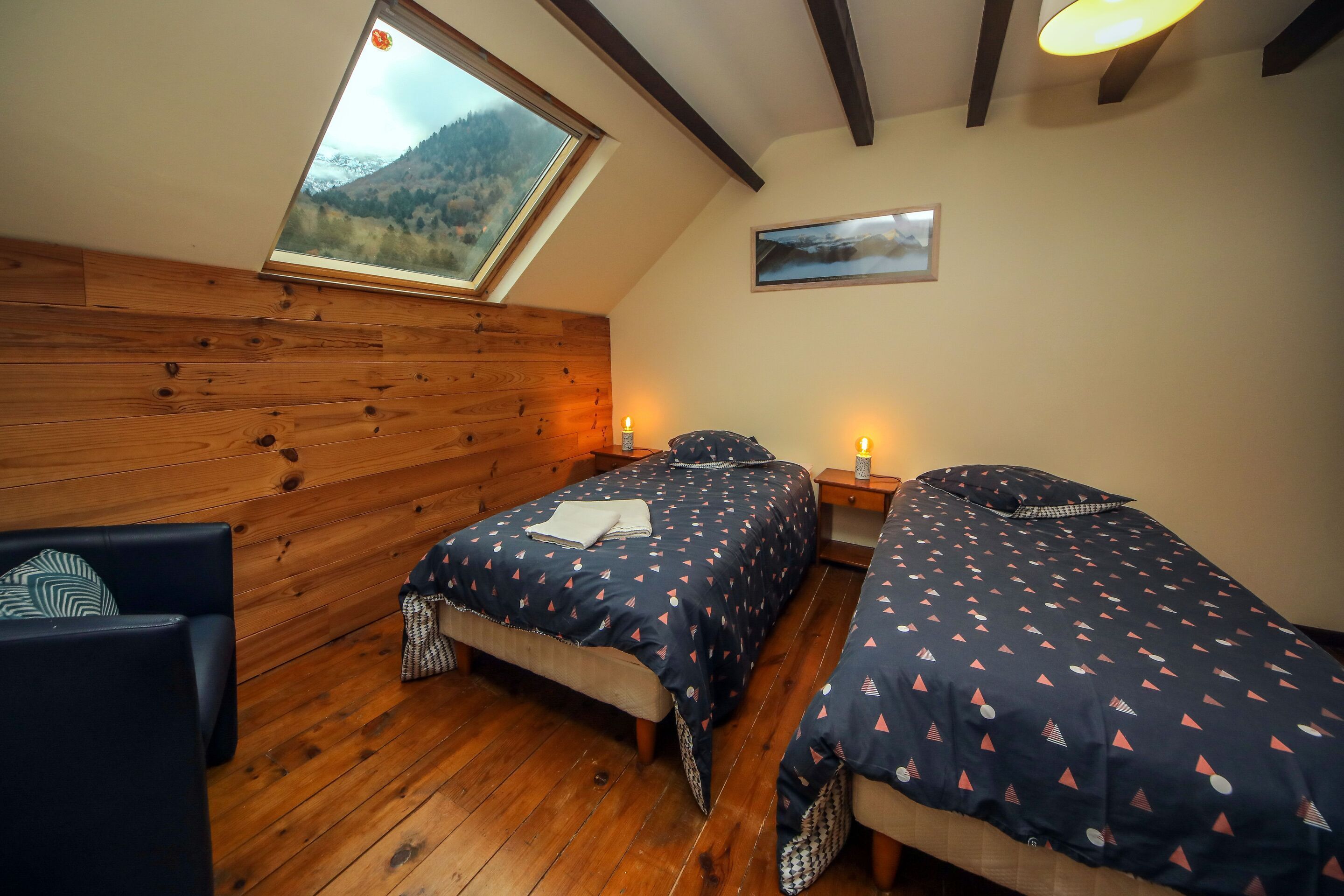 Una habitación acogedora con paredes de madera, un tragaluz, dos camas con ropa de cama estampada, una silla y una mesita de noche con lámpara.