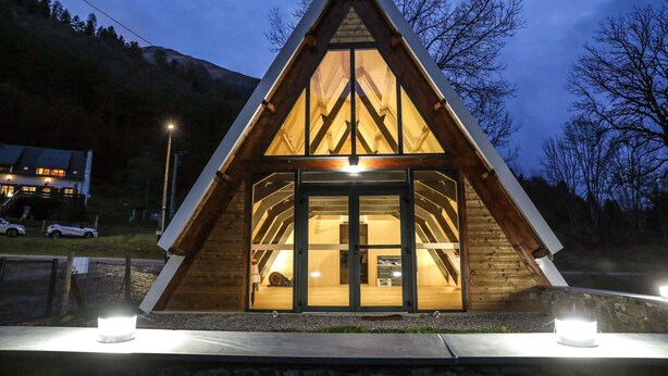 Una cabaña de madera triangular con entrada de puerta de cristal, rodeada por una base de piedra e iluminada por dos luces en el suelo.