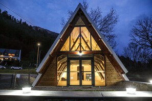 A triangular wooden cabin with a glass door entrance, surrounded by a stone base and lit by two ground lights. - Le Refuge de L'isard (Bagnères-de-Bigorre)