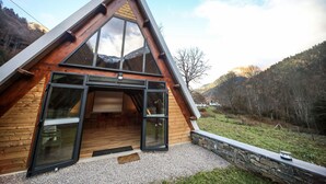 Una cabaña moderna con techo triangular, ventanas amplias, pared de piedra y vista a una ladera boscosa.