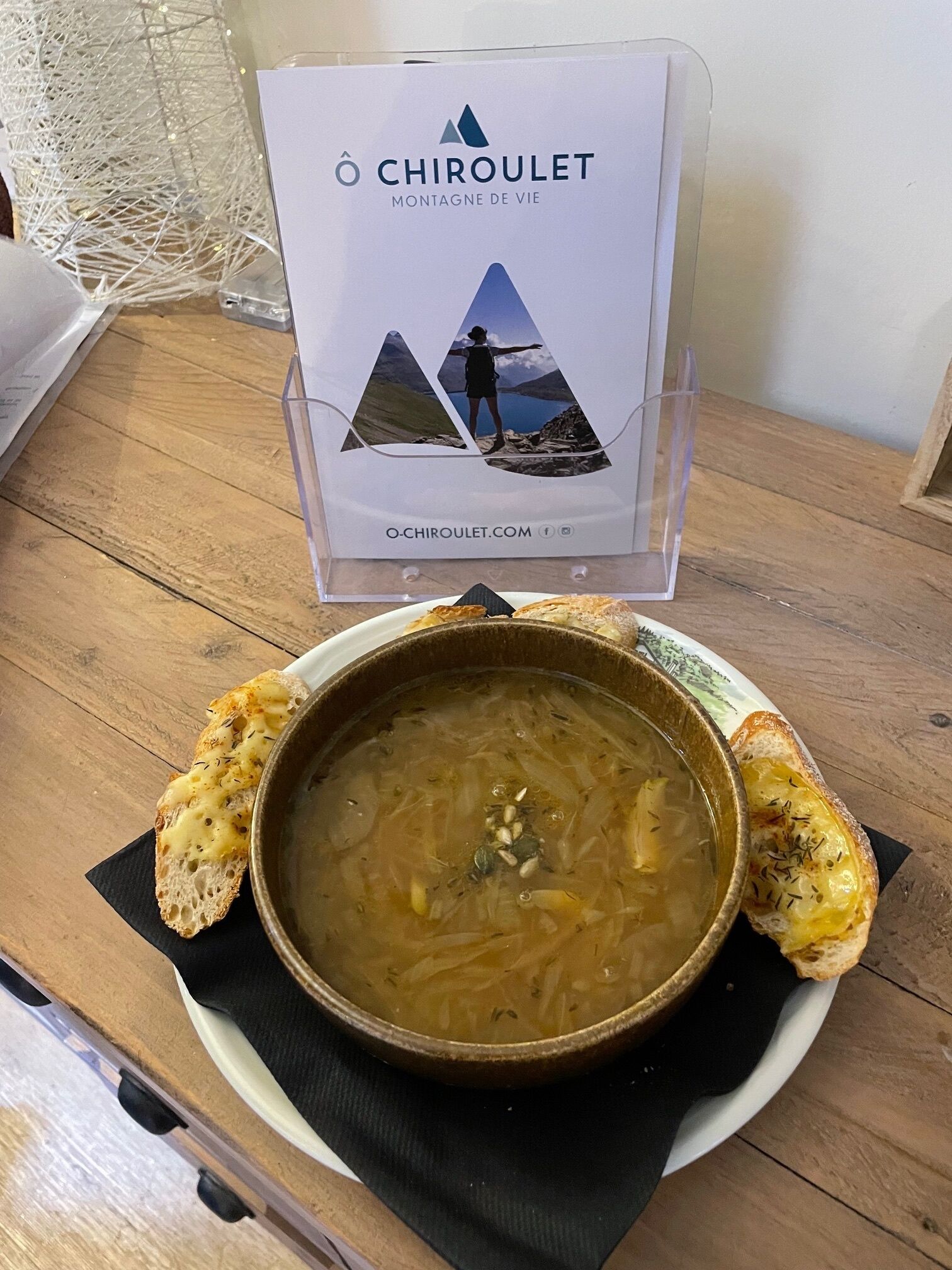 A wooden table with a bowl of soup and toasted bread, a sign with 'CHIROULET MONTAGNE DE VIE' and a website URL, and a decorative string of lights in the background.