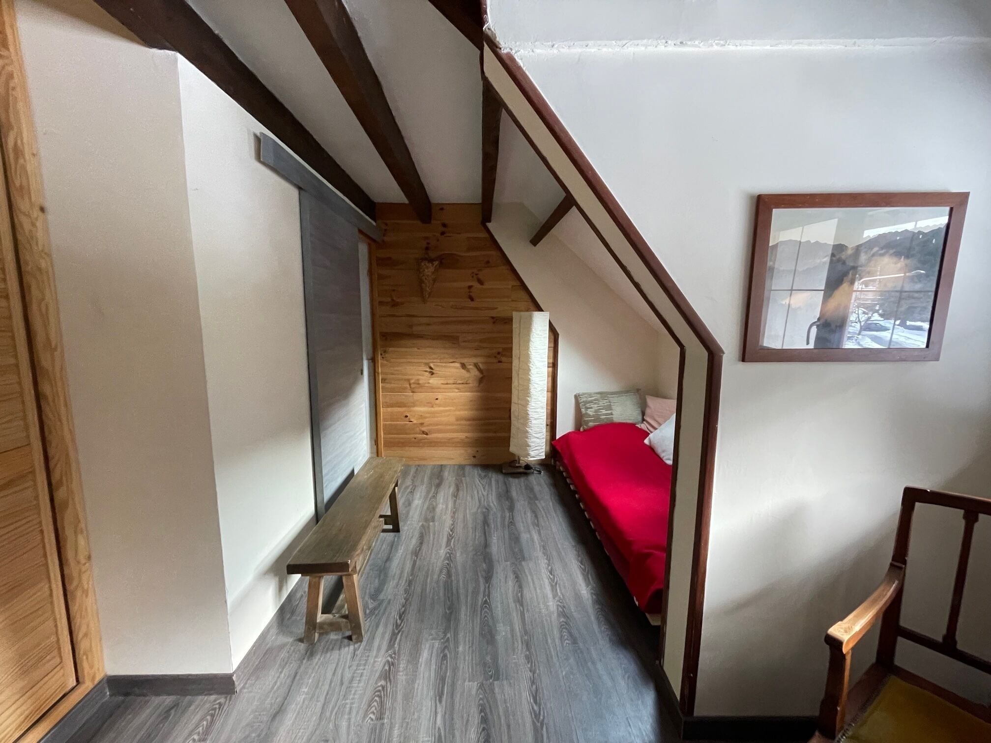 Un estrecho pasillo con suelo de madera, un banco, una cama con manta roja y un cuadro en la pared.