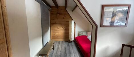 Un estrecho pasillo con suelo de madera, un banco, una cama con manta roja y un cuadro en la pared.