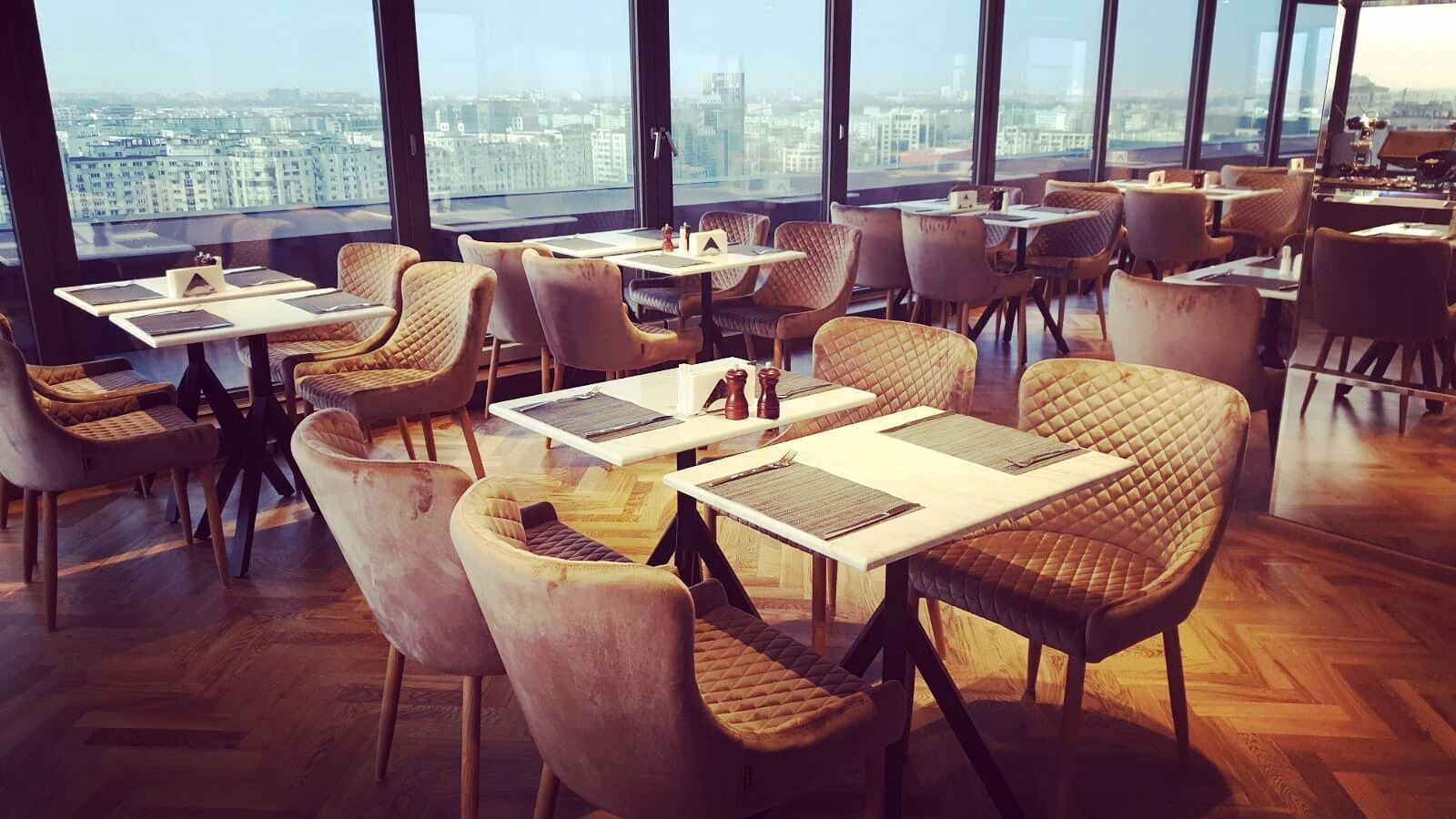 A modern dining area with high tables and cushioned chairs, large windows offering a city view.