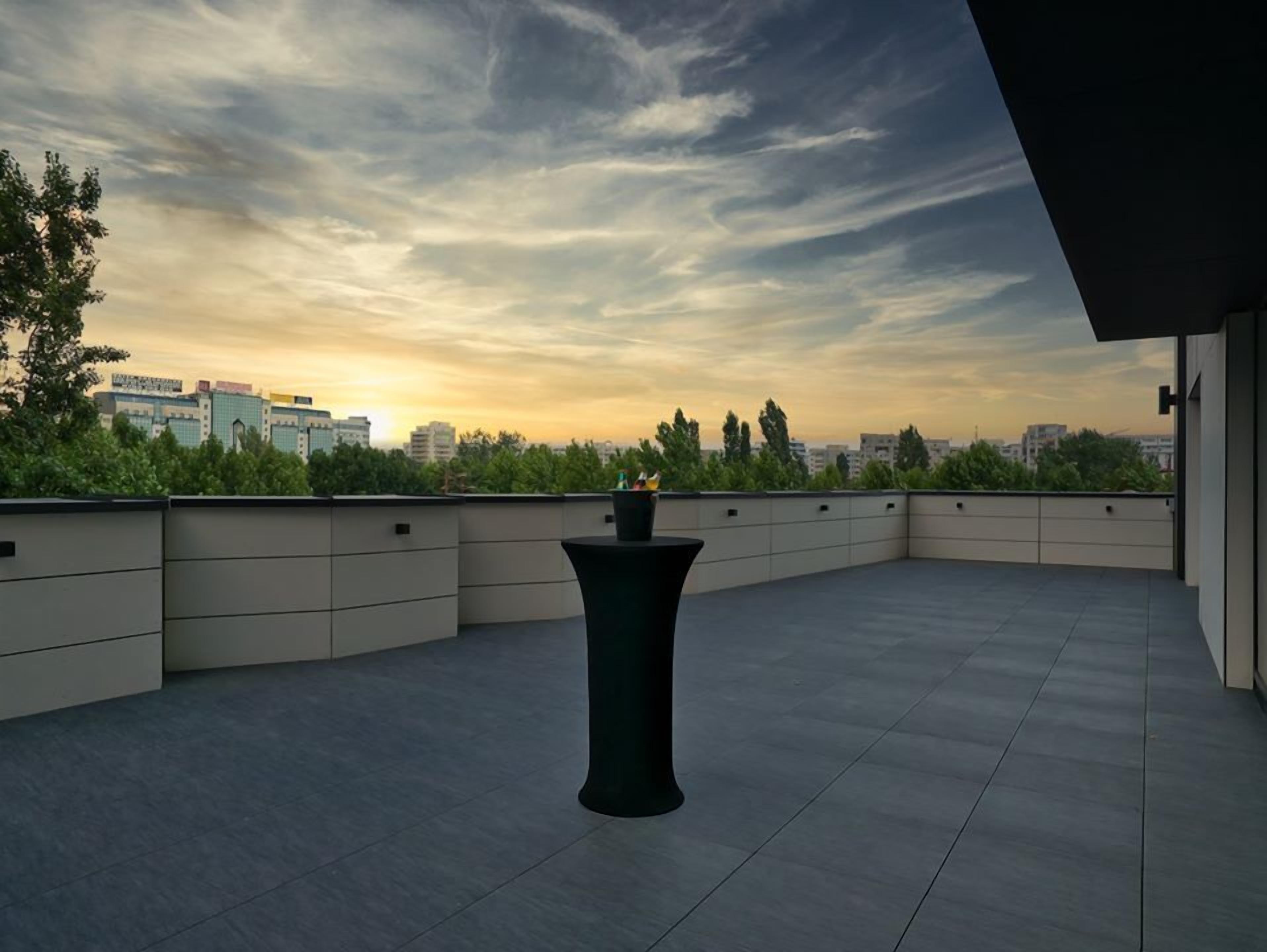 Een dakterras met een zwarte tafel en stoelen, omringd door een lage muur en een uitzicht op de stad bij zonsondergang.
