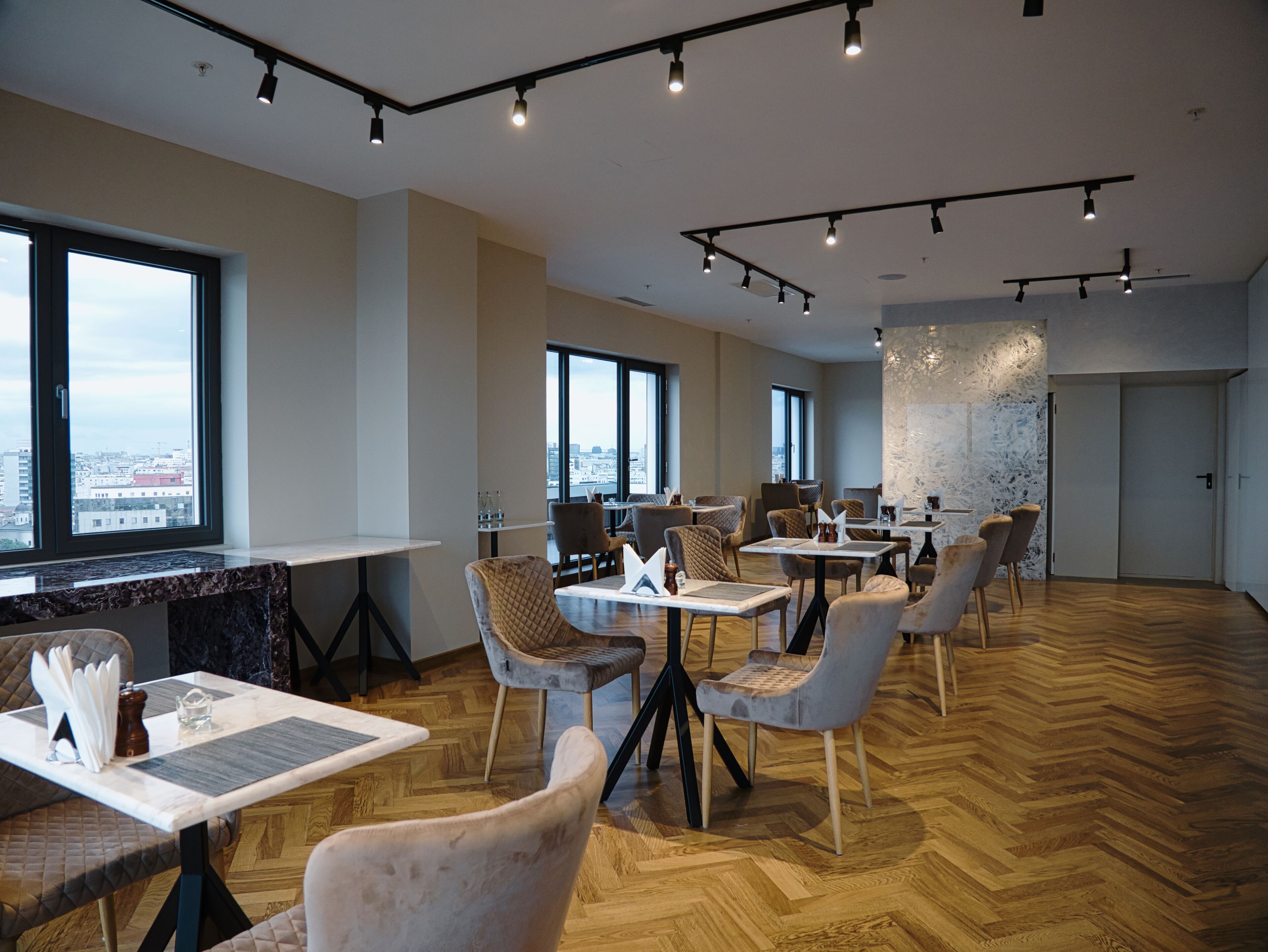 Un comedor moderno con varias mesas y sillas, ventanas amplias que ofrecen vistas a la ciudad y un suelo de madera en espiga.