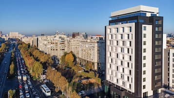 A modern hotel with a prominent glass facade, situated next to a busy road with trees and parked cars.