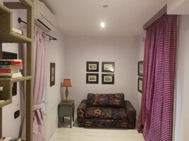 A living room with a sofa, a small table with a lamp, a bookshelf, and a wall with framed pictures.
