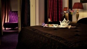 Una habitación de hotel con una cama, un sillón púrpura, una cortina roja y una mesa con una lámpara y flores.