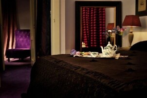 A hotel room with a bed, a purple armchair, a red curtain, and a table with a lamp and flowers.