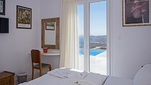 A hotel room with a bed, a chair, a small table, and a window with a view of the sea. - Chrysanthemo Villas (Mykonos)