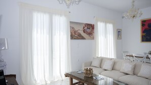 A living room with a glass coffee table, beige sofa, white curtains, and a chandelier. - Chrysanthemo Villas (Mykonos)