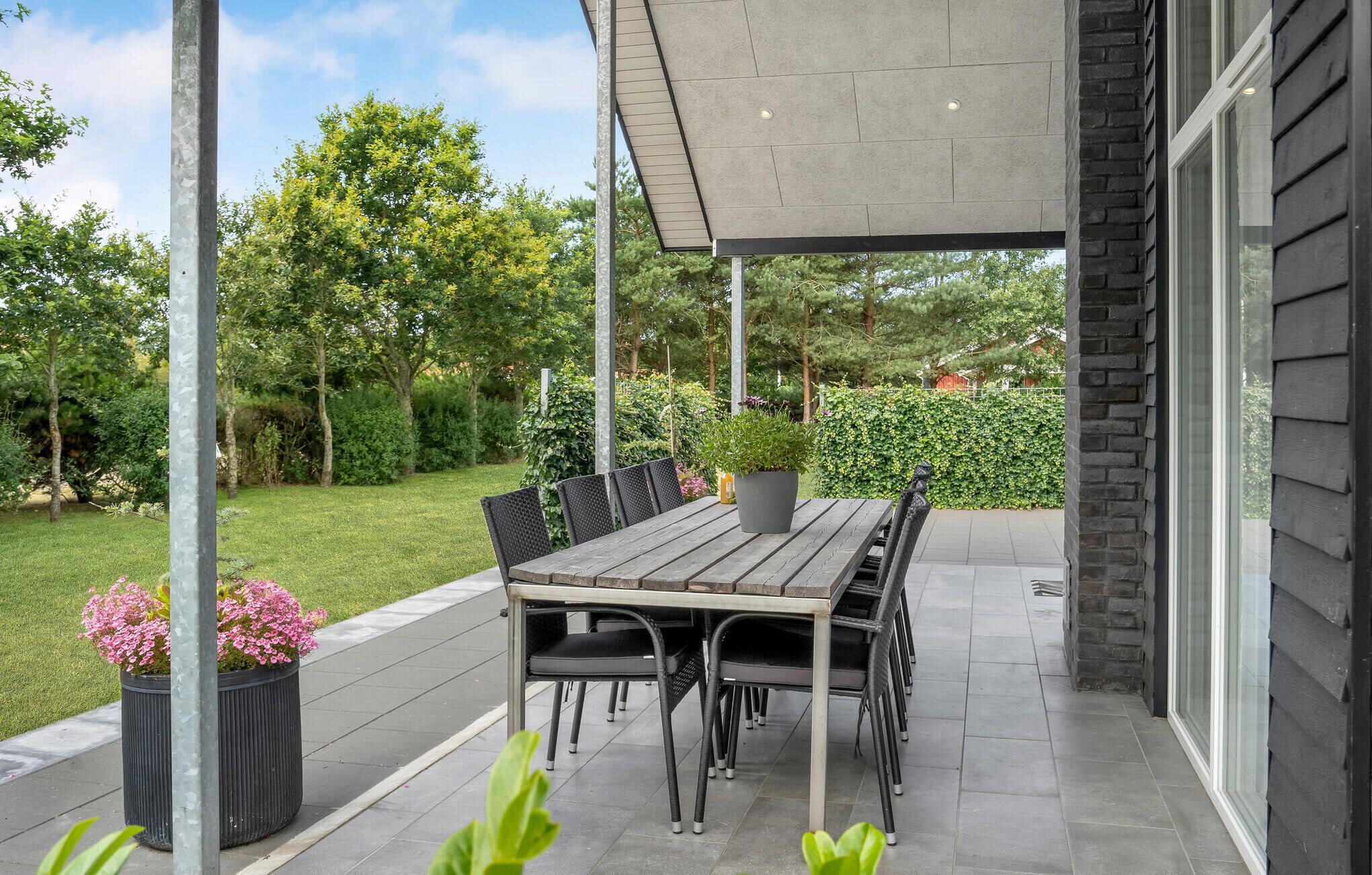 Une terrasse couverte avec une table et des chaises en bois, entourée de verdure et d’une haie.