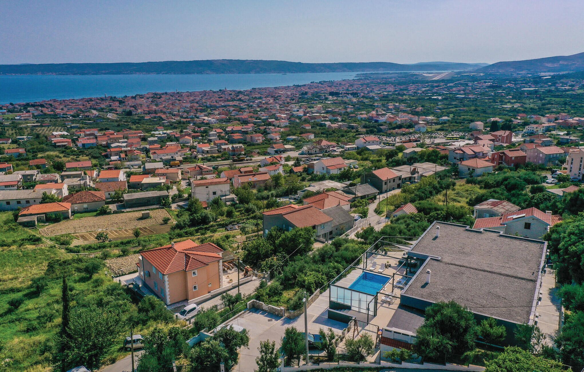 Vista aèria d'una població costanera amb cases de teulada vermella, una extensa massa d'aigua i un edifici modern amb piscina.
