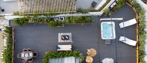 An aerial view of a rooftop patio with a hot tub, lounge chairs, a dining table, and surrounding greenery.