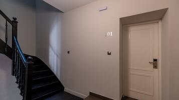 Una escalera con barandillas negras que lleva a un rellano, una puerta blanca con el número 101, y un interruptor de luz en la pared.