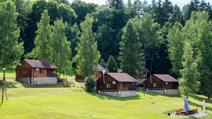A row of wooden cabins with stone foundations, surrounded by a grassy field and dense forest. - Zloty Potok Resort (Lesna)