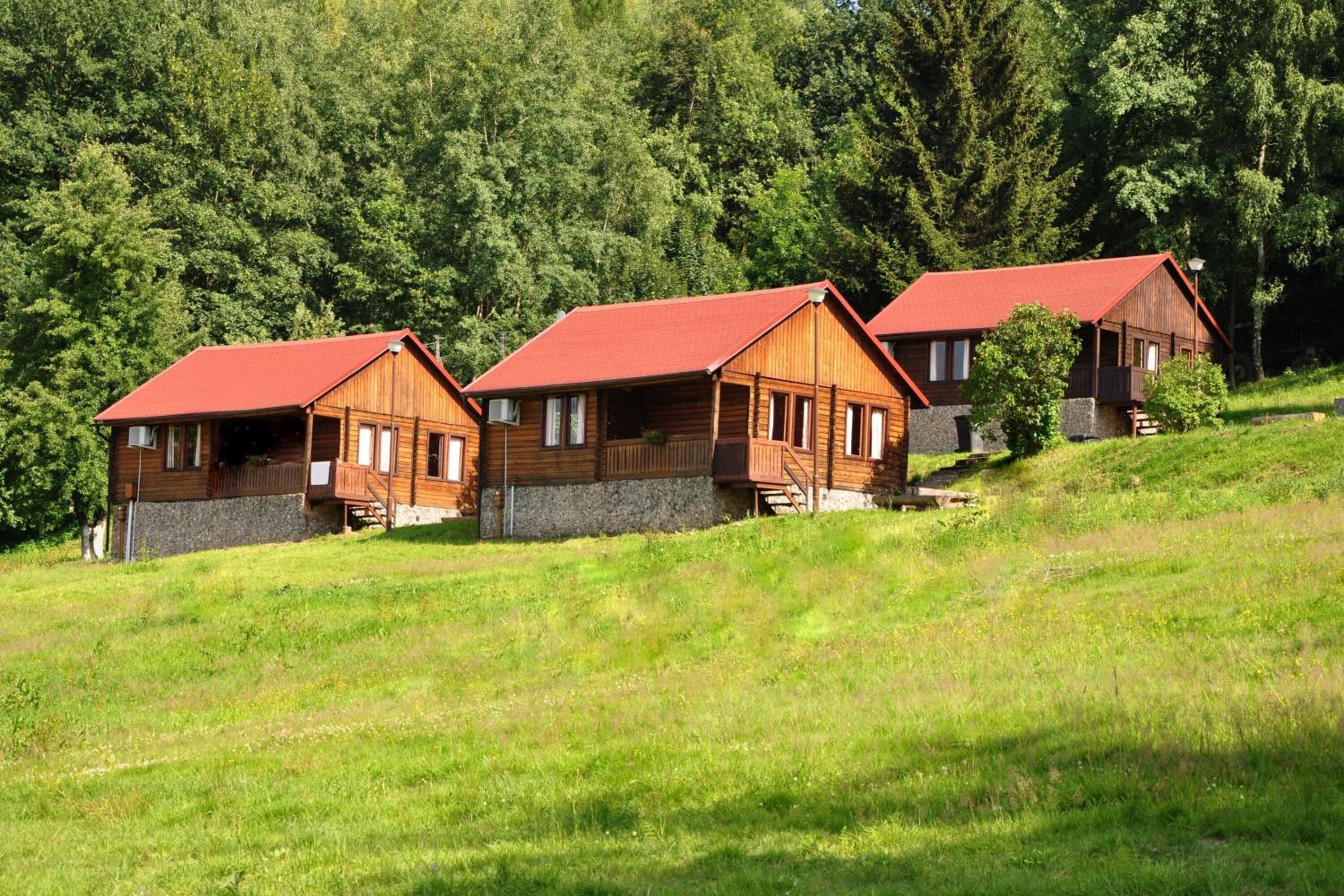 Drei Holzhütten mit roten Dächern auf einer grasbewachsenen Hügelkuppe, umgeben von dichtem Wald.