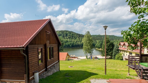 A wooden cabin with a red roof overlooking a lake, surrounded by lush greenery and a picnic table. - Zloty Potok Resort (Lesna)
