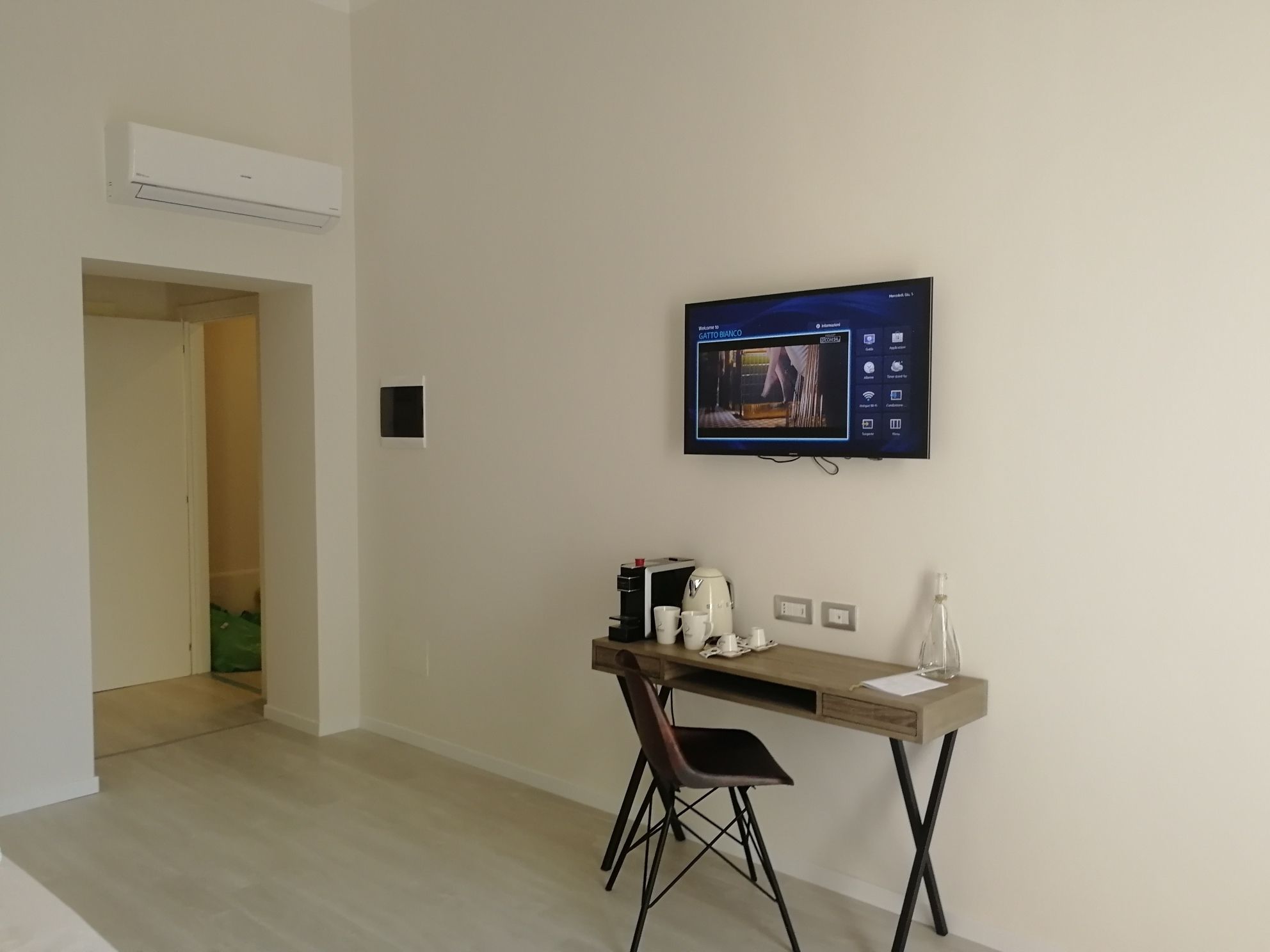 Una habitación moderna con televisión de pantalla plana en la pared, un escritorio de madera con una silla, y una pequeña mesa con cafetera y tazas.