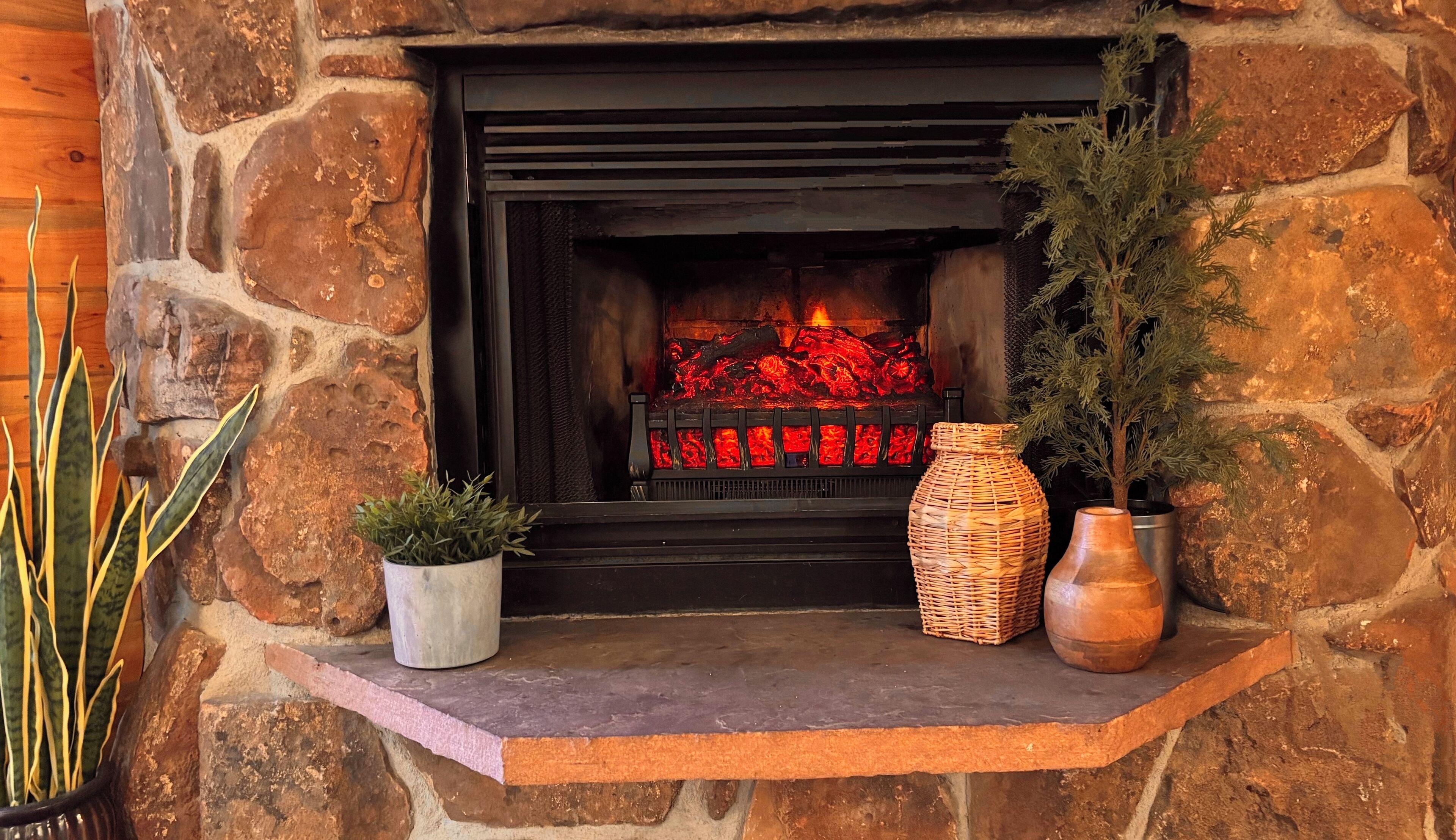 Une cheminée en pierre avec un feu, une petite étagère avec une plante en pot, un panier tressé et un pot en argile.