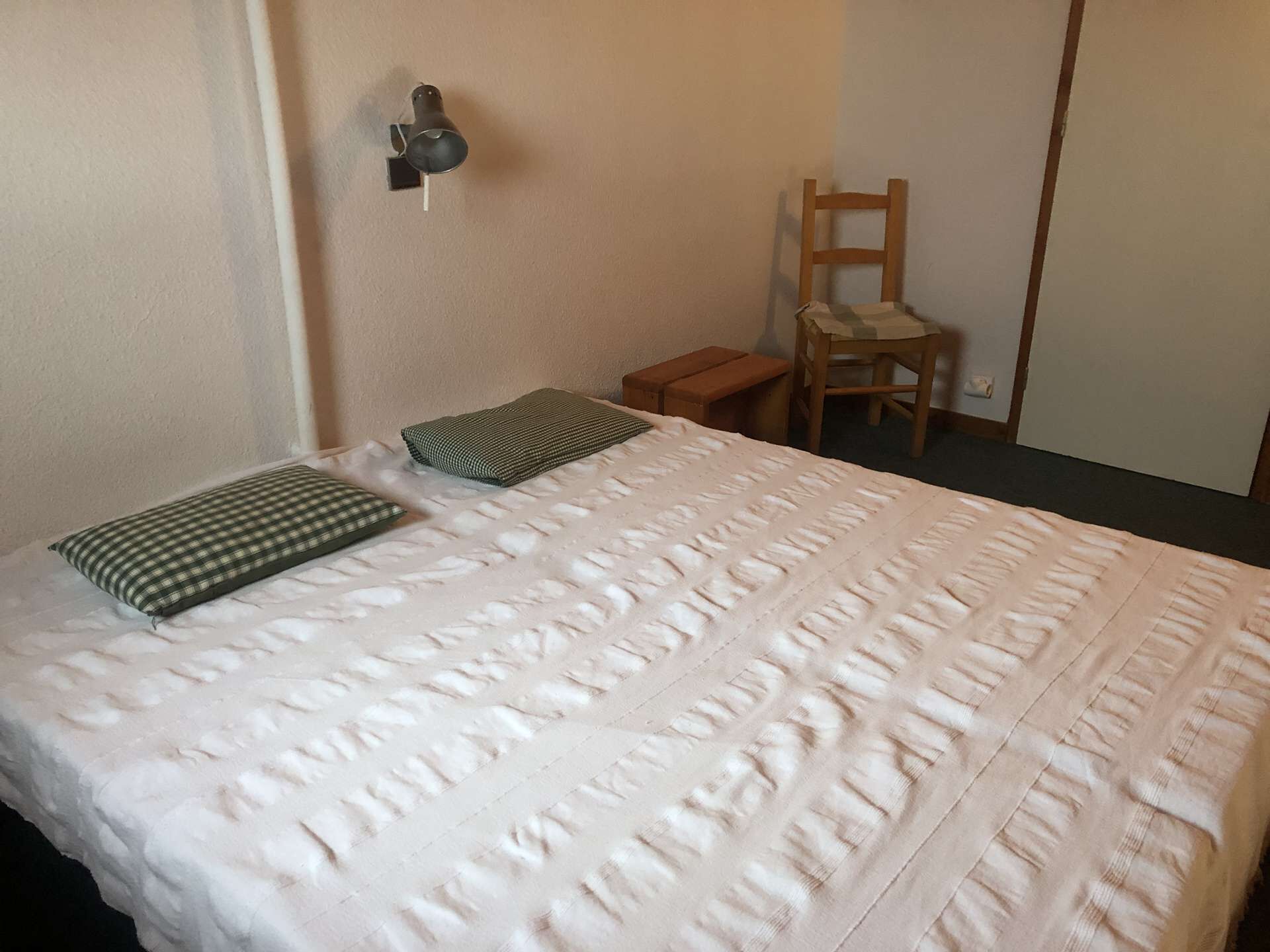 A neatly made bed with a checkered pillow and a quilted blanket, a small wooden stool, and a chair against the wall.