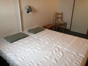 Un lit bien fait, avec un oreiller à carreaux et une couverture matelassée, un petit tabouret en bois et une chaise adossée au mur.