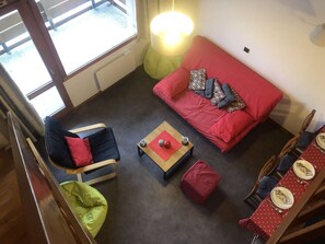 A cozy living room with a red sofa, a blue armchair, a wooden coffee table, and a dining area with a red tablecloth.