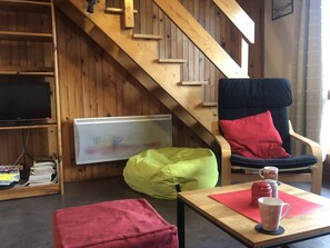 Une pièce chaleureuse avec un escalier en bois, une télévision, un fauteuil poire, une chaise avec des coussins et une table avec une tasse.